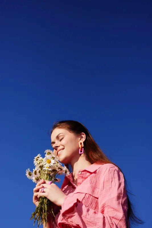 Young woman with long hair in a pink shirt holding daisies against a clear blue sky, smiling joyfully, embodying the essence of spring and happiness