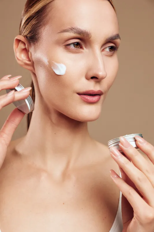Young woman applies moisturizing cream on face closeup natural skincare routine with clean skin and neutral beige background, beauty care concept with healthy complexion.