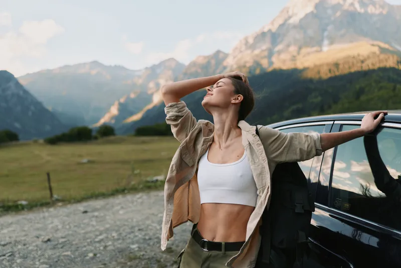 One woman with brunette hair leans on a car in a mountain valley, wearing a white crop top and beige jacket, tilting her head back in a free, relaxed moment.