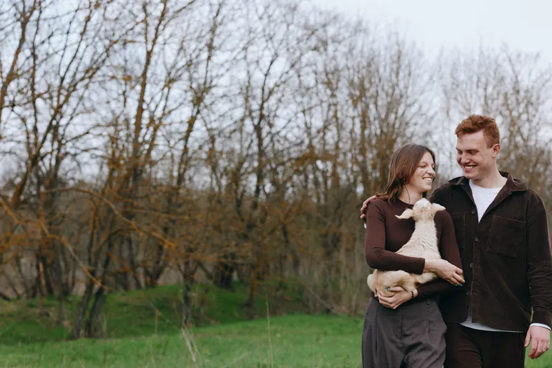 Happy couple outdoors with a dog, smiling and affectionate, walking through a park in bright daylight, enjoying nature and each other, expressing warmth and companionship on a calm day.