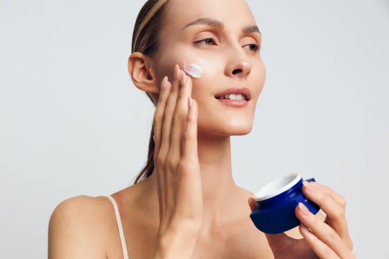 Young woman applying white face cream on cheek with hand. Close-up portrait on light background. Concept of skincare, beauty, wellness, and daily self-care routine. Natural lighting.