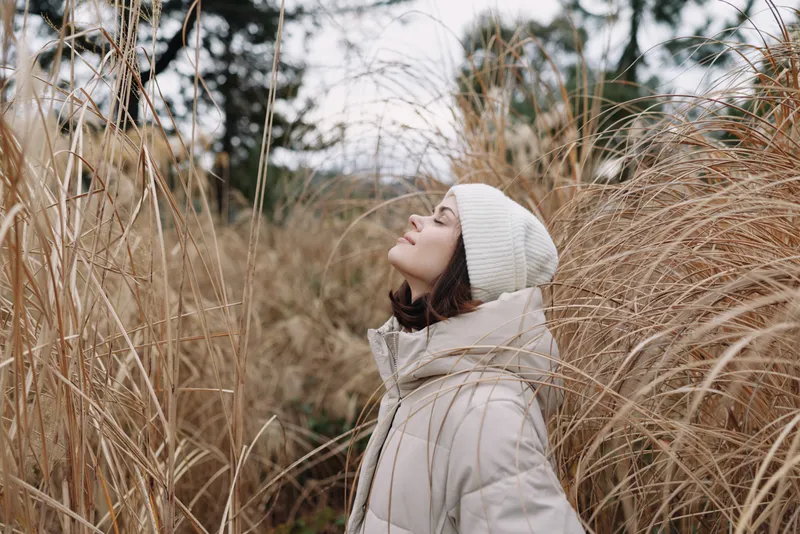 Woman in a warm beige coat and white beanie stands among tall dried grasses, gazing upward with a calm, reflective expression in a quiet nature field.