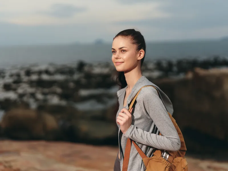 Young woman hiking by the sea, wearing a gray hoodie and backpack, smiling with a serene expression, showcasing adventure and nature