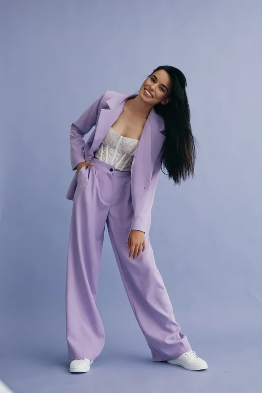 Young woman wearing lilac pantsuit and white sneakers posing stylishly against a soft purple background, showing modern fashion and relaxed confident attitude.