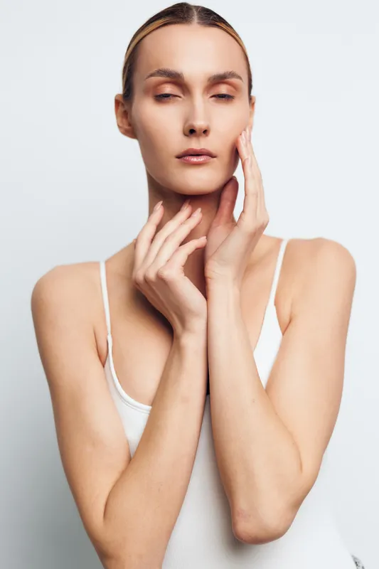Young woman with clear skin posing with closed eyes and hands touching her face and neck on white background. Concept of skincare, beauty, wellness, and natural health care.