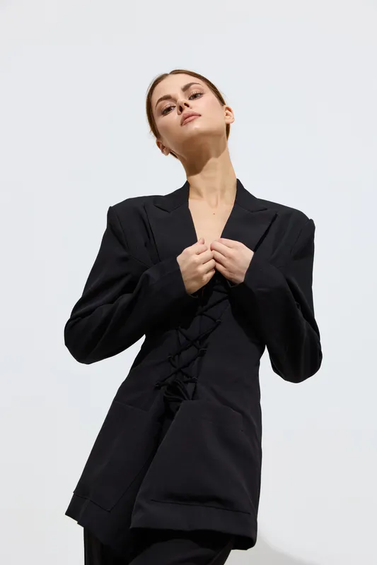 Fashion woman posing confidently in a black tailored blazer with strong lines against a plain bright background, showcasing minimalistic style and modern elegance.