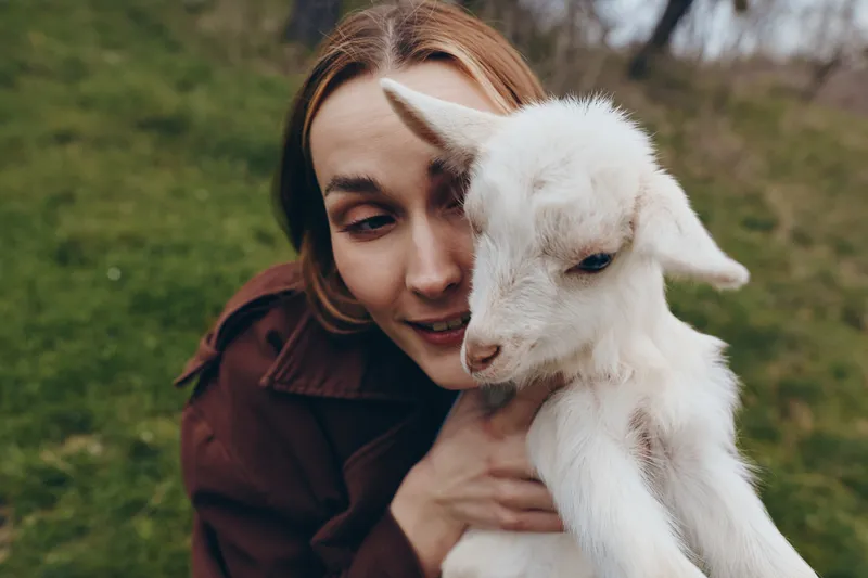 Soft close up of a smiling woman holding a young goat kid in her arms, outdoors on green grass. Warm, tender moment conveying companionship, nature, and gentle care in soft daylight.