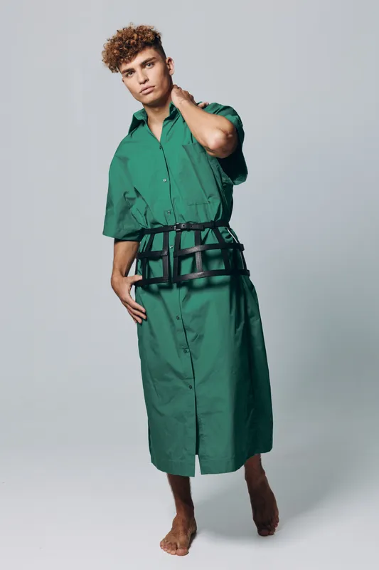 Young man with curly hair wearing long green shirt dress and black belt posing barefoot on gray background. Fashion model with casual style and relaxed posture in studio lighting.