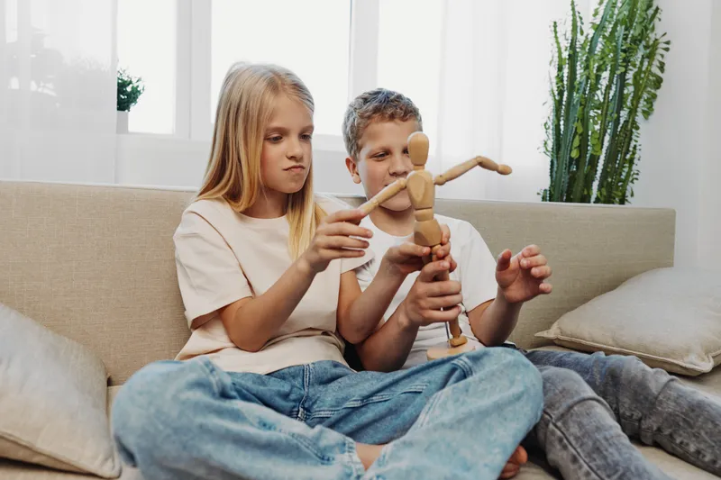 Two children sitting on a sofa playing with a wooden mannequin figure in a bright living room. Concept of childhood creativity, play, and sibling interaction at home.