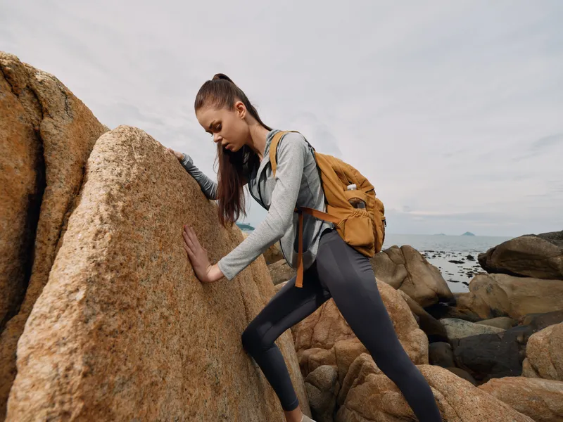 Young woman climbing rocks outdoors, showcasing determination and strength, wearing sporty attire and carrying a backpack in a natural setting