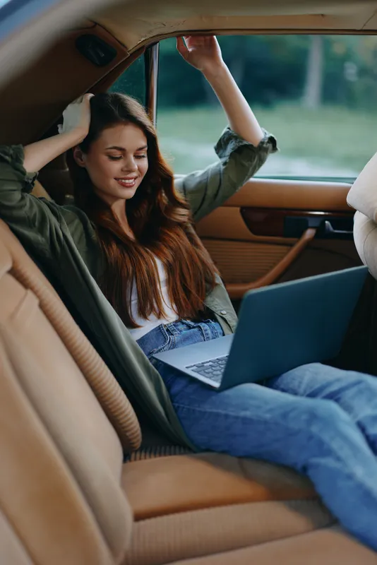 Young woman relaxing in a car while working on a laptop, showcasing a casual outfit, joyful expression, outdoor background, and warm color palette.