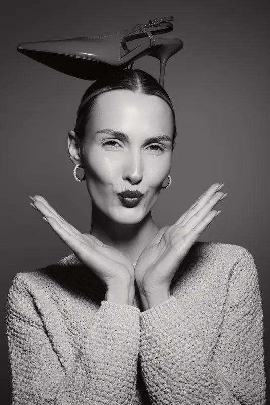 Portrait of a smiling white caucasian woman with pale skin wearing a knitted sweater and hoop earrings posing with a high heel shoe on her head in black and white.