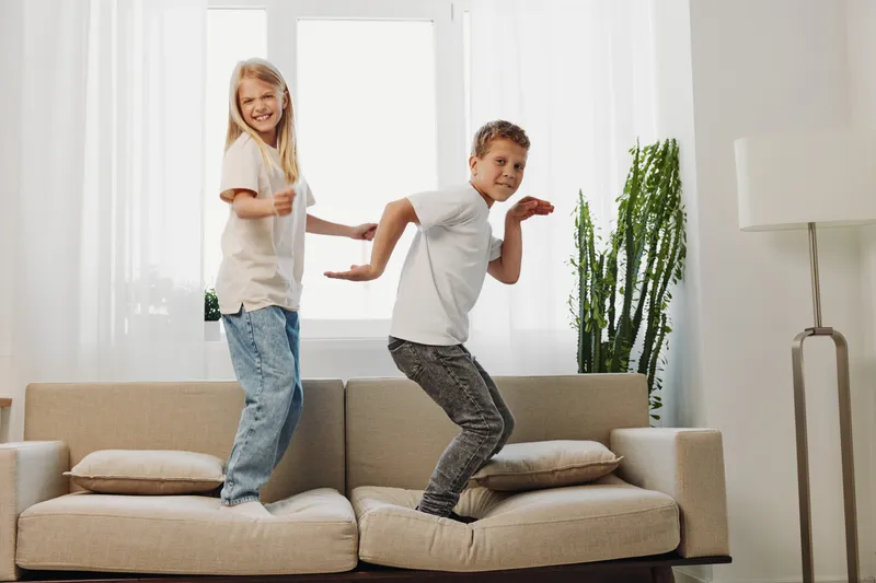 Happy children jumping on beige sofa in bright living room. Boy and girl having fun, playful mood, natural light. Concept of family joy, childhood energy, and indoor activity.