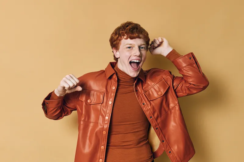 Young man with curly red hair wearing leather jacket and turtleneck celebrating excitedly with raised fists on beige background in studio. Happiness and energetic expression.
