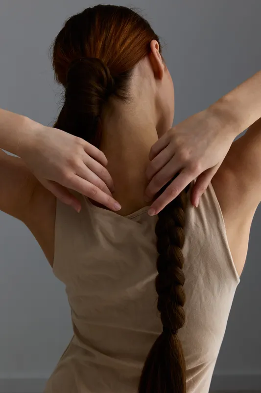 Woman with long braided hair touching neck and shoulders wearing beige tank top against plain gray background, natural light emphasizing skin texture and calm pose.