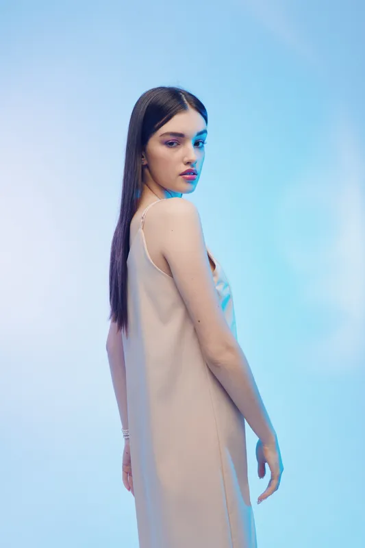 Young woman with long dark hair wearing a beige dress poses sideways on a blue gradient background. Concept of woman beauty fashion with soft studio lighting and elegant style.