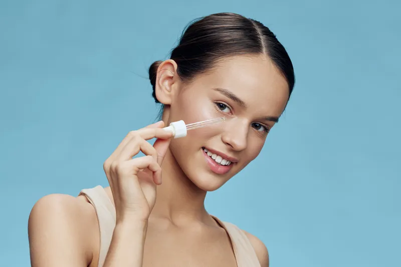 Young model applying skincare serum with a dropper against a light blue background, showcasing healthy skin and a fresh look