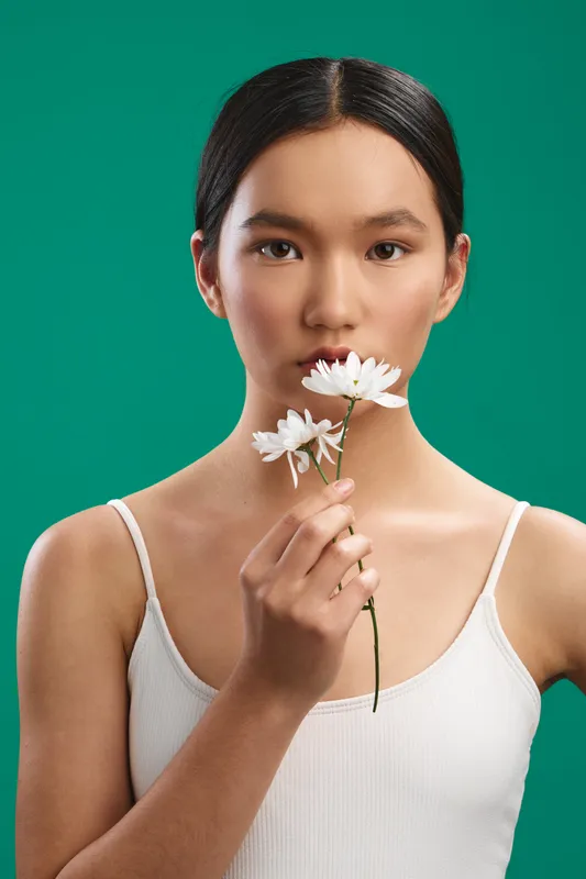 Asian woman stands against a teal backdrop, holding a white flower near her lips, a calm and intimate portrait that exudes simplicity and elegance.