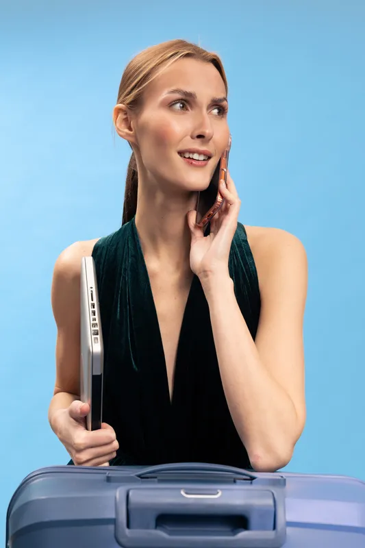 Young woman holding laptop and talking on phone with suitcase in front of her. Concept of business travel, communication, and professional lifestyle on light blue background.
