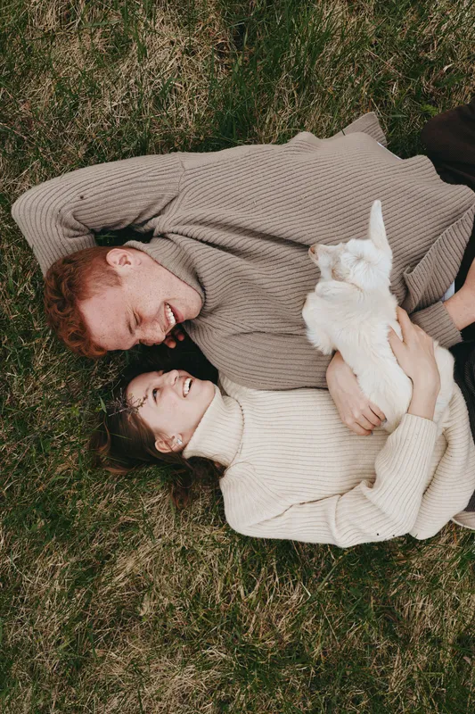 Lifestyle shot of a smiling couple lying on grass, cuddling a playful goat kid, warm sweaters, autumn light, intimate outdoor moment, cozy nature mood, gentle romance