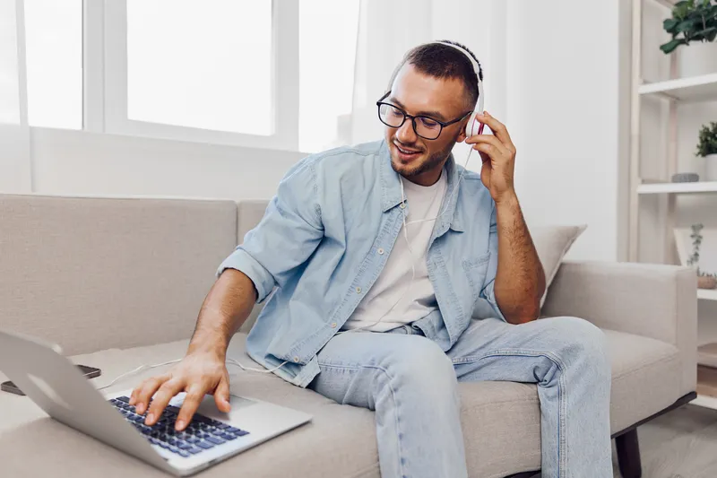 Casual man working on a laptop while listening to music, wearing headphones and denim clothing, in a bright modern living space, showcasing productivity and relaxation.