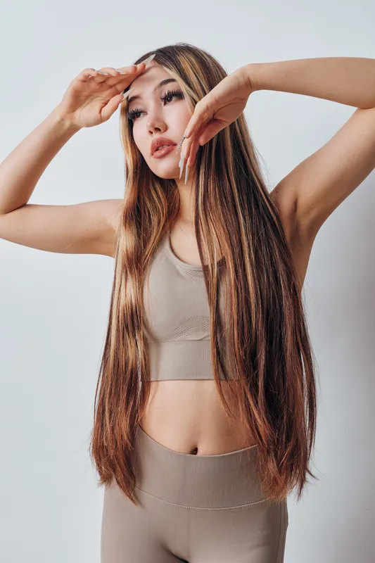 Young woman with long hair posing with hands near forehead in neutral outfit against white background. Female model showing trendy hairstyle and casual fashion indoors.