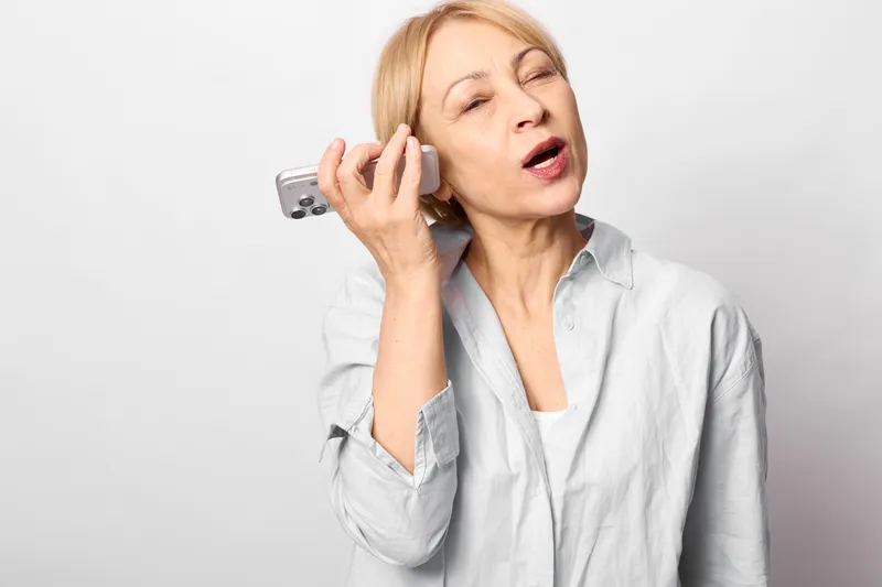Senior woman holding smartphone to ear, making a call in communication moment, eyes closed as if feeling headache, showing stress, isolated on light gray background