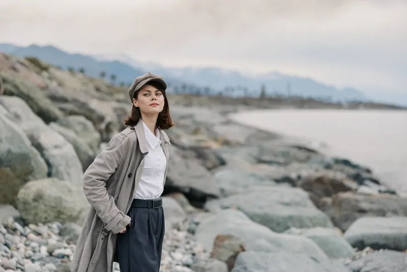 Young woman in a long coat and cap stands on rocky beach near calm sea with mountains in the background. Overcast sky creates soft natural light and peaceful mood.
