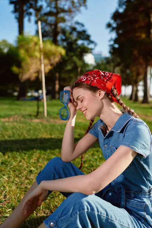 smiling woman relaxing outdoors with red bandana and sunglasses sitting on grass in sunny park on summer day