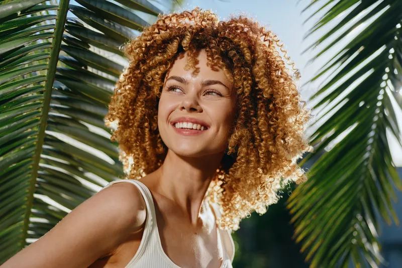 smiling woman with curly hair enjoying tropical sunlight near green palm leaves, wearing white tank top with natural summer vibes and radiant joyful expression.