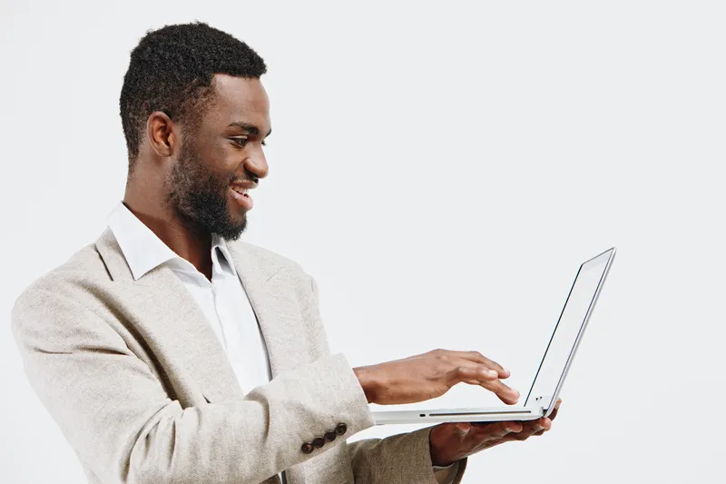 Confident man in beige sweater and white shirt using laptop computer, standing against plain white background, performing work or online communication, modern lifestyle concept.