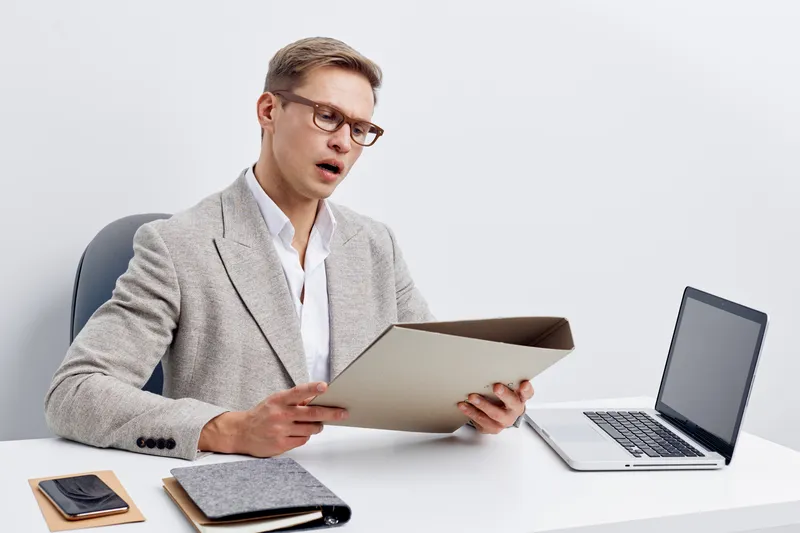 Professional man in business attire reading documents while sitting at a desk with a laptop, smartphone, and notebooks in a modern office setting. Business, work, professional, concentration, desk.