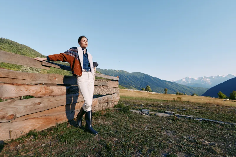 Woman stands by wooden fence in mountain meadow at sunrise, wearing jacket, scarf and rubber boots. Landscape view with rolling hills and clear blue sky for outdoors leisure.