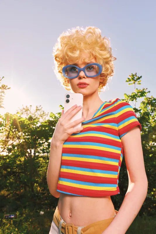 A stylish woman in a colorful striped crop top and blue sunglasses stands outdoors in bright sunlight, conveying retro fashion, carefree mood, and vintage glamour.
