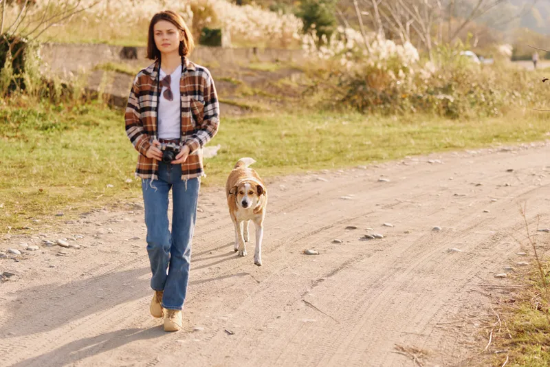 Young girl walking a dog along a dusty countryside path, wearing a plaid shirt and jeans, enjoying a sunny day, travel vibe and casual exploration with a loyal companion.