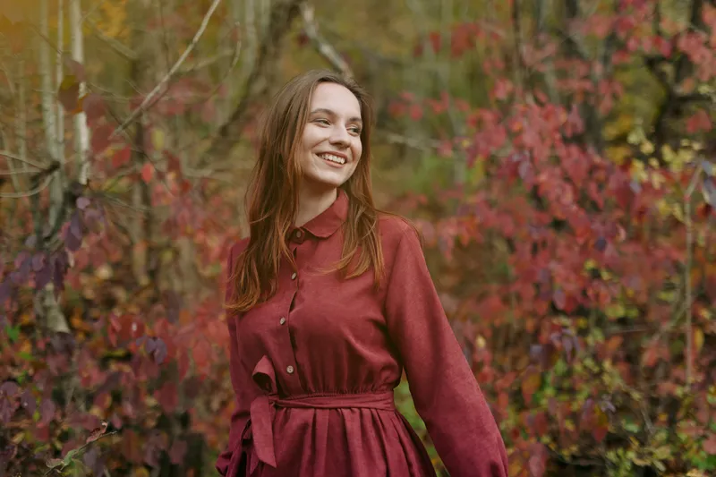 A woman in a burgundy dress stands amidst autumn foliage, presenting an authentic, credible portrait with natural light and a candid, warm smile.