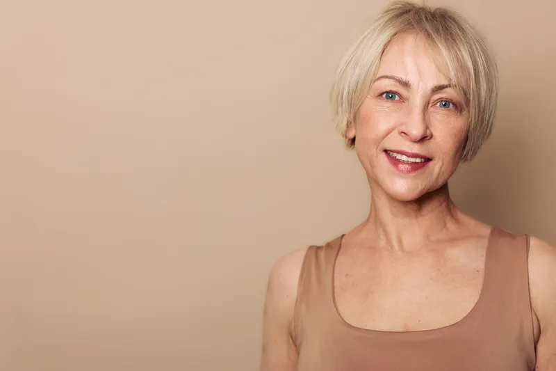 Smiling mature woman with short blonde hair in beige top poses against neutral background. Concept of natural beauty, aging gracefully, confidence, and positive lifestyle for women.