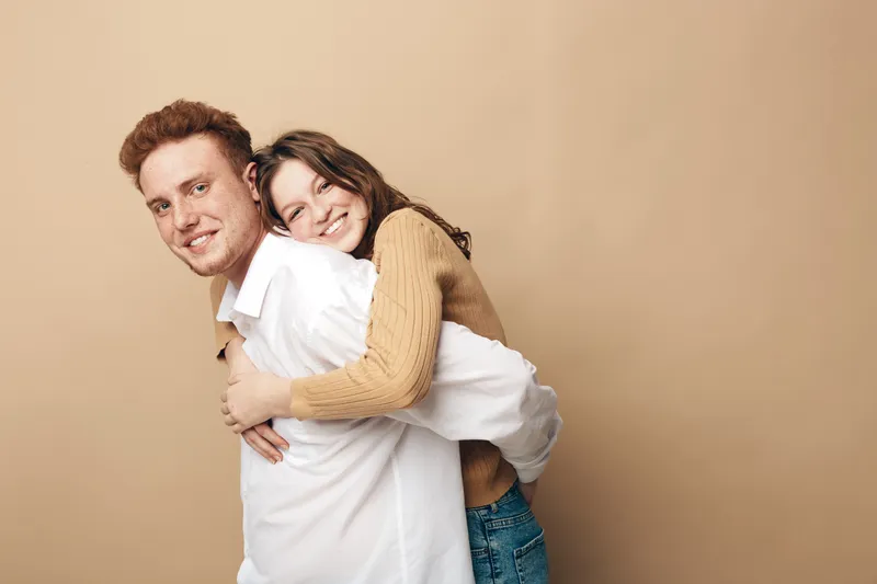 Smiling young couple in casual clothes embraces against beige background. Happy man and woman enjoy playful moment together. Concept of love, friendship, and relationship.