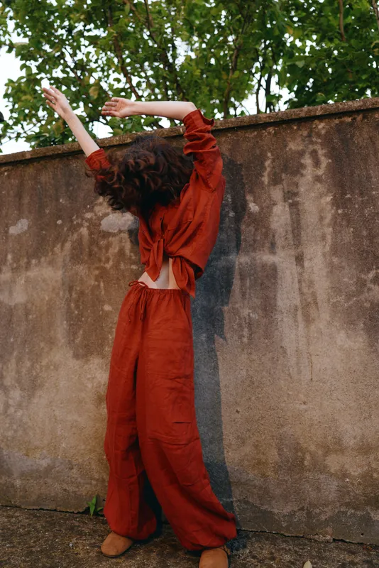 Back view of a woman in a rust red jumpsuit outdoors, arms raised in a relaxed pose, natural light, weathered wall, candid fashion moment