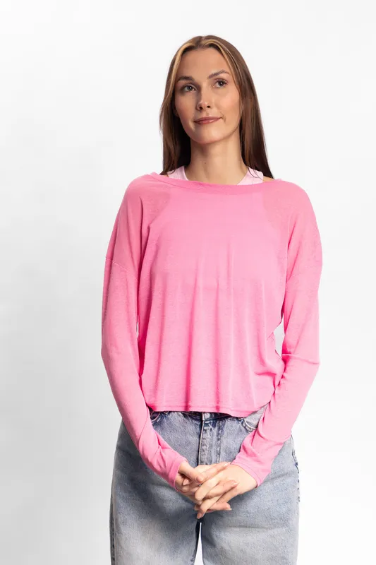 Young woman with long hair wearing pink sweater and denim jeans stands against white background. Casual fashion look with relaxed pose and thoughtful facial expression.