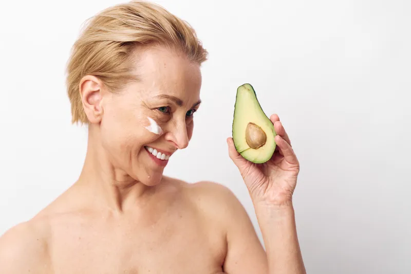 Smiling mature woman with short hair holds half of avocado near face, applying natural cream on cheek. Concept of skincare, healthy lifestyle, natural beauty, and organic cosmetics.