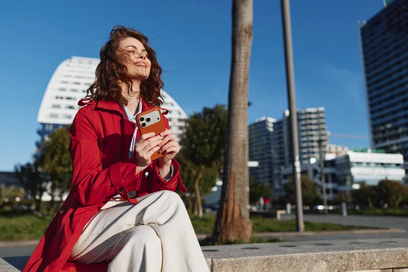 Smiling woman with curly hair wearing a red jacket using a smartphone outdoors. Urban park setting with tall buildings, bright sunlight, casual lifestyle moment.