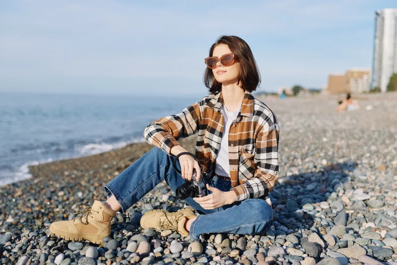 Woman sits on a rocky beach in casual attire with a plaid shirt, jeans and boots, gazing toward the sea under bright sunshine, conveying a relaxed outdoor lifestyle moment for travel or leisure