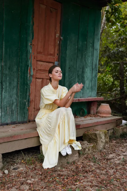 Woman portrait on rustic porch in long yellow dress sitting with hands clasped in nature authentic inclusive mood quiet meditation and relaxation