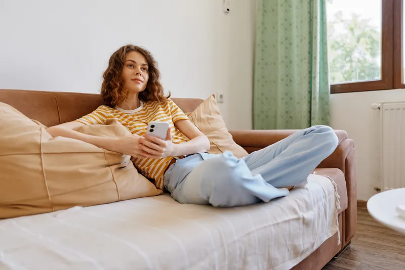 A young woman with curly brown hair relaxes on a beige sofa at home, browsing a smartphone, casual mood and cozy atmosphere.