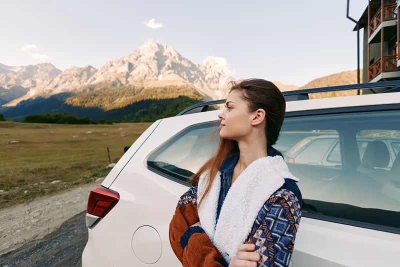 Woman leaning against white car at roadside with mountains backdrop, travel roadtrip landscape in autumn wearing patterned jacket, portrait of relaxed traveler enjoying scenic nature view.
