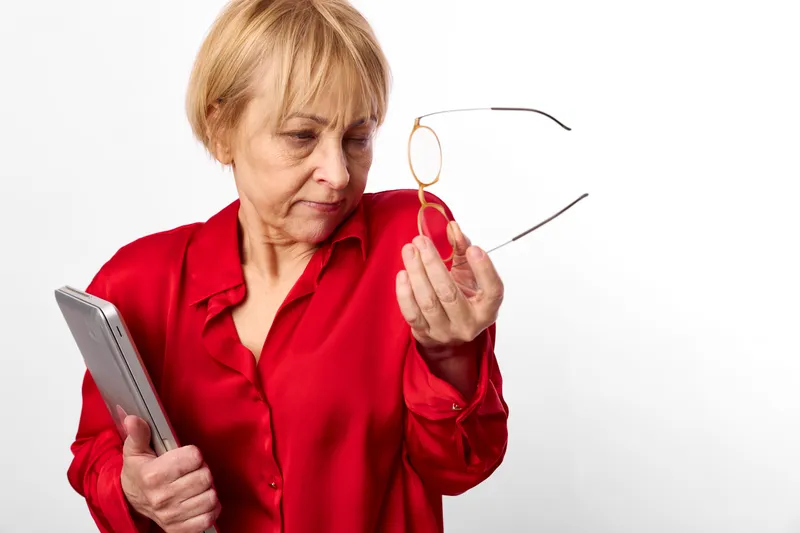 Elderly woman holding glasses near her face, trying reading a book, concerned expression with reduced eyesight, isolated on bright white background, closeup aging vision concept