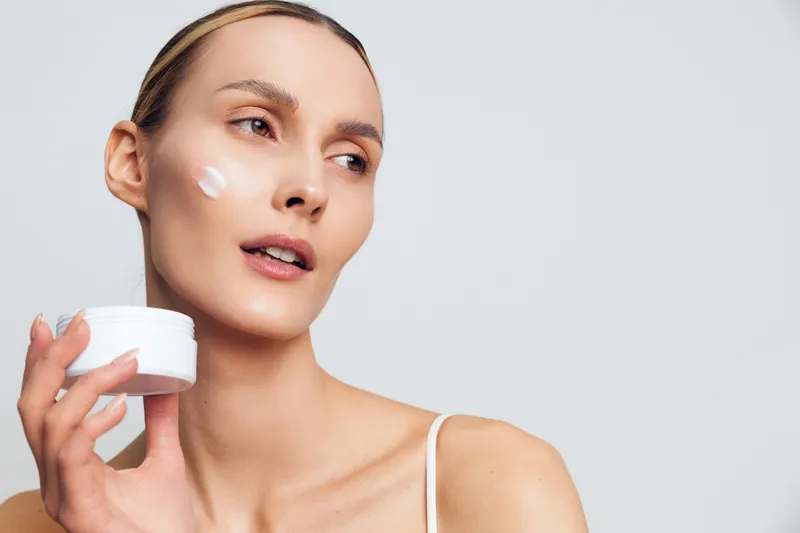 Young woman holding white cream jar applying moisturizer on clean face soft natural light smooth skin beauty skincare routine minimal background