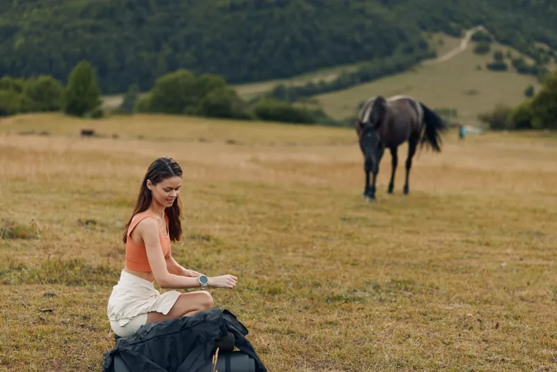 A young woman sits on a bag in a sunny countryside meadow, texting on a smartwatch, while a horse grazes in the distance; a calm, candid outdoor moment.