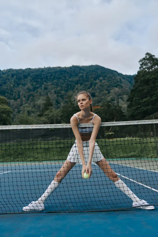 Female tennis player in sportswear poses on a court, exuding determination and focus against a serene nature backdrop